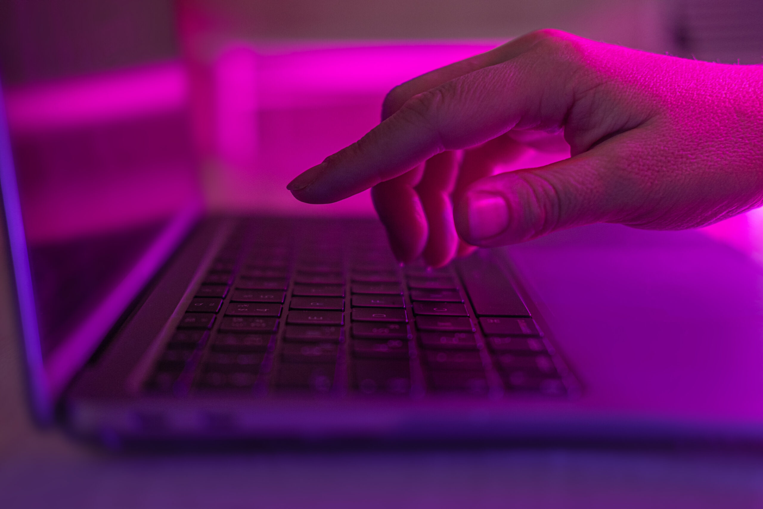 Hand hovers over laptop keyboard, illuminated by purple light.